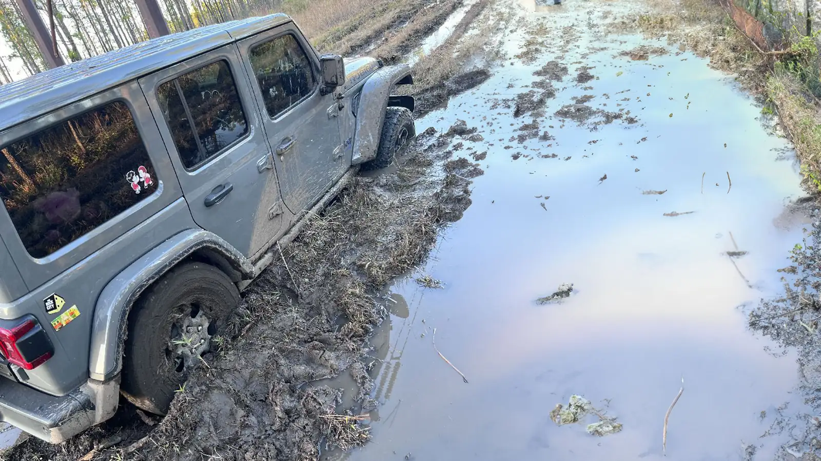 A four-wheel-drive vehicle sunk to its axles in mud beside a flooded, unpaved track in a forest