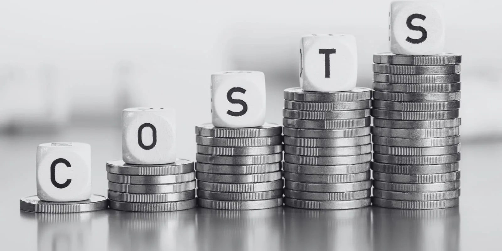Stacks of coins ascending in height with dice spelling COSTS balanced on top, representing the real and rising costs of software development